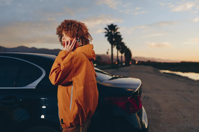 Woman lifestyle outdoors near car at sunset, hooded jacket in warm orange, standing on road with distant mountains, one hand touching hair, calm travel mood, soft cinematic light today