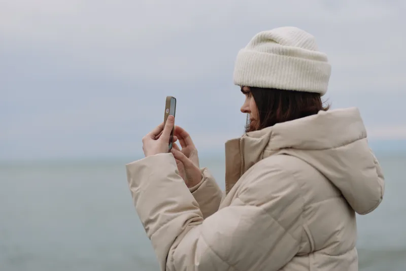Outdoor photography of a woman in a warm jacket and beanie, capturing scenery on a smartphone by the sea. Casual winter mood, cloudy sky, calm water and distant horizon, reflecting a tranquil coastal