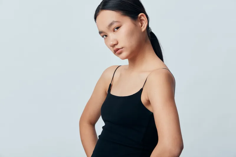 Young woman with long dark hair in ponytail wearing black sleeveless top posing with confident look on plain background. Portrait of female model with natural makeup and casual style.