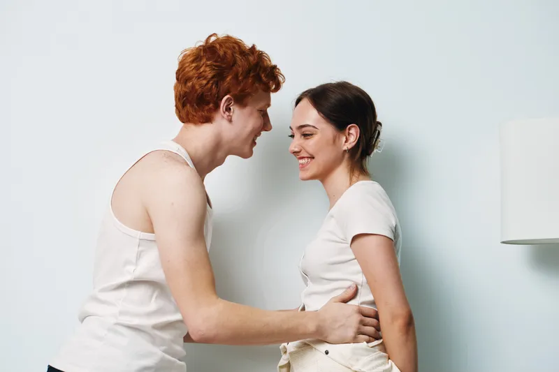 Young couple with red hair and dark hair smiling and embracing indoors, showing affection and happiness in casual white clothing on light background.