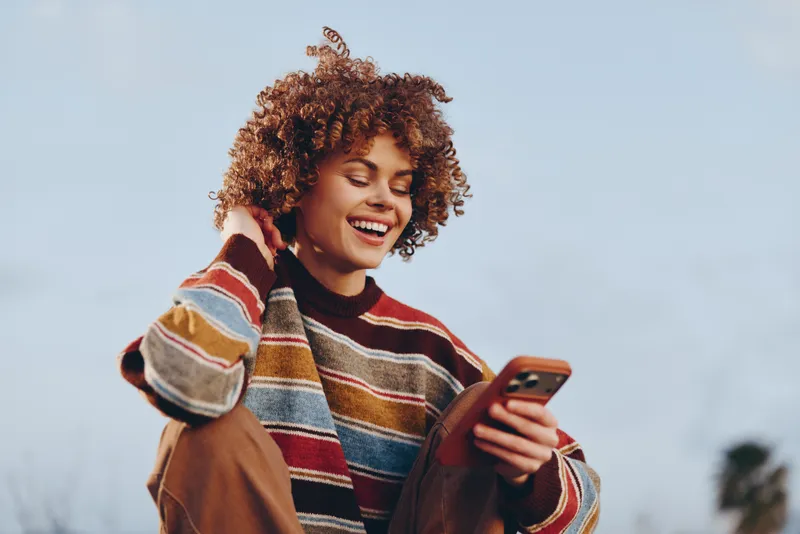 Woman lifestyle outdoors, smiling with curly hair, holding smartphone, casual striped sweater, candid communication moment, sunlight, relaxed posture, friendly expression, clear sky