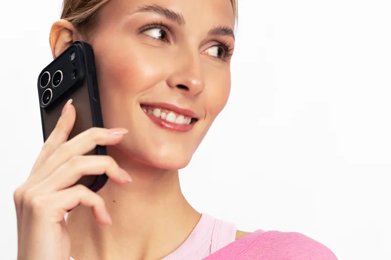 Smiling young woman holds smartphone to ear with hand, looking aside on white background. Concept of communication, technology, happiness, and modern lifestyle in casual wear.