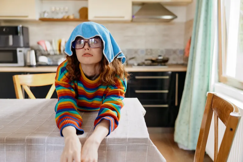 Young girl in a striped rainbow sweater sits at a table in a bright kitchen with a blue towel on her head, reaching forward with a curious, focused expression.