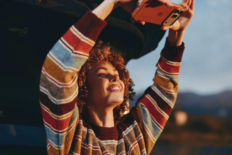 Woman lifestyle portrait outdoors, curly hair and smiling face as she holds a smartphone for selfie at sunset, arms raised in casual autumn sweater, soft natural light, scenic sky