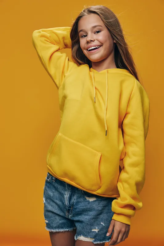 Teenage girl smiling and posing in yellow hoodie and denim shorts on yellow background, casual style with long hair and joyful expression, studio portrait with bright colors.