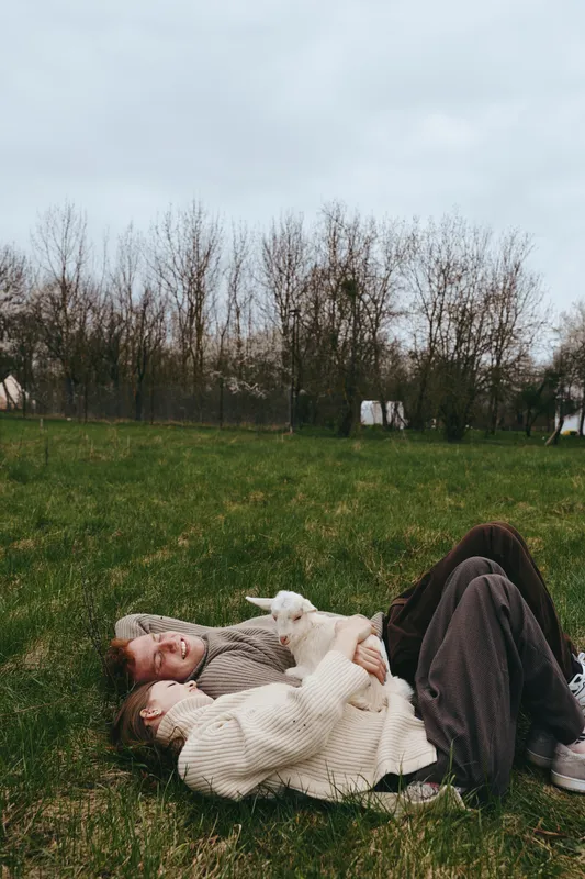 Lifestyle outdoor scene with a couple relaxing on a grassy field, wearing chunky sweaters, enjoying calm moments together as a curious goat kid grazes nearby under soft overcast light.