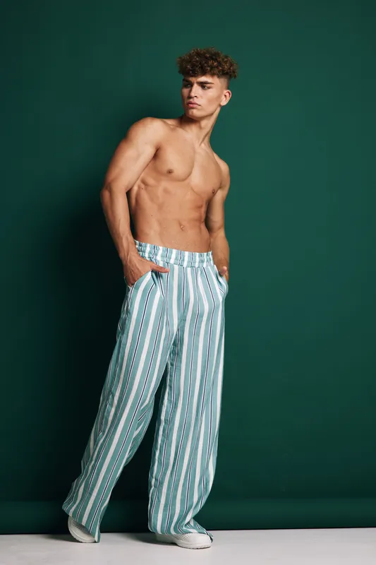 Young man with curly hair posing shirtless in striped wide pants and white sneakers against green background. Fashion, casual style and confident expression in studio setting.