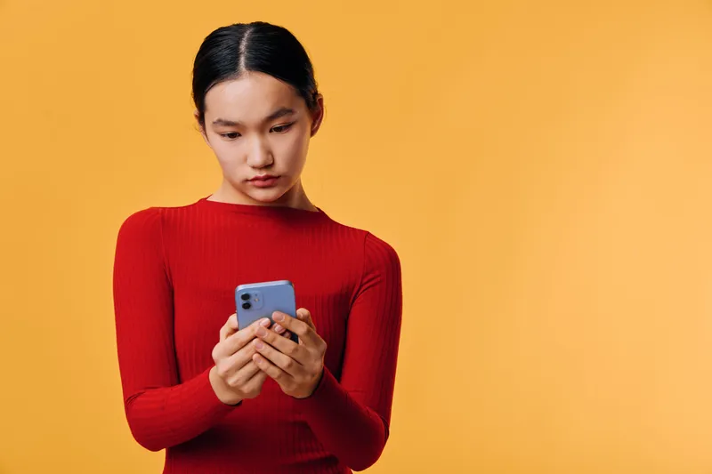 Young woman with dark hair in red sweater looking at smartphone with focused expression on orange background. Technology, communication, mobile device and concentration concept.