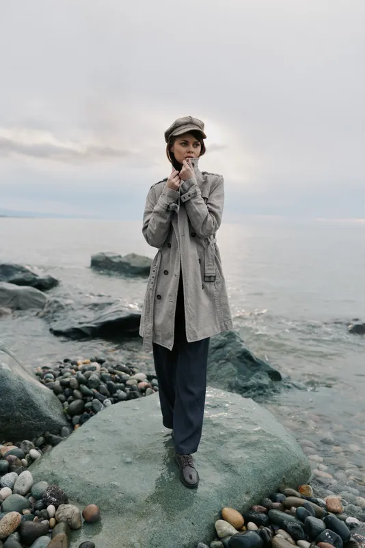Young woman in a grey coat and hat stands on rocky shore near calm sea under cloudy sky. Moody outdoor portrait with natural light, fashion and autumn concept by the water.