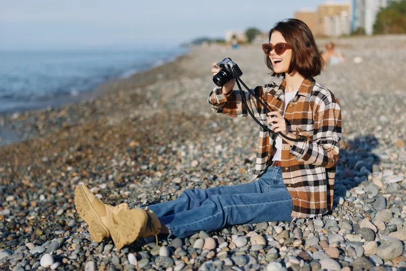 Photographer sitting on a pebbled beach with a camera, wearing a plaid shirt and denim, enjoying daylight by the sea, capturing a casual outdoor portrait amid coastline scenery and ocean breeze
