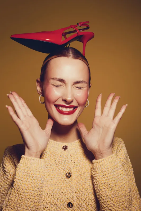 Smiling woman with closed eyes wears red high heel shoe on head, shows hands near face. Studio portrait on brown background. Concept of fashion, fun, and playful mood.