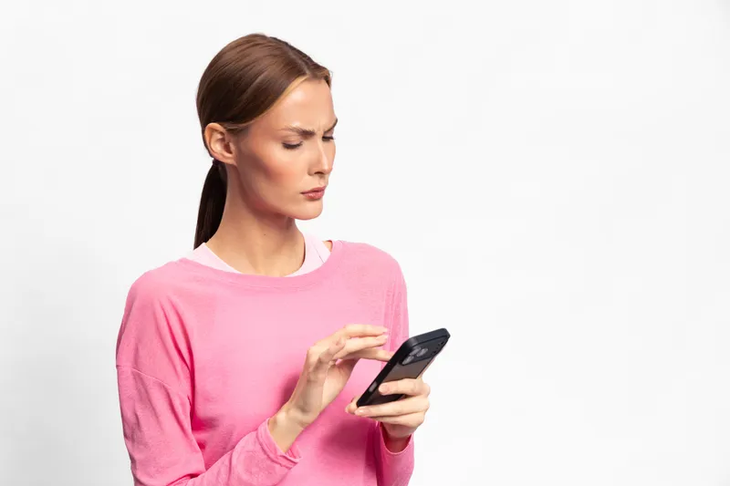 Young woman in pink sweater looks confused while using smartphone on white background. Concept for technology frustration, communication problems, and digital confusion.