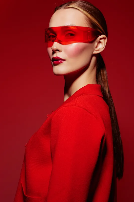 Stylish woman with long hair wears red jacket and futuristic red visor glasses on red background. Concept of fashion, modern style, and bold color contrast.