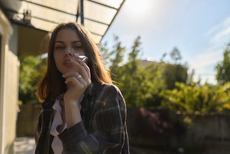 A casual woman outdoors on a sunny day, lighting a cigarette, captured in a candid lifestyle moment. The portrait highlights personal routines, relaxation, and urban leisure