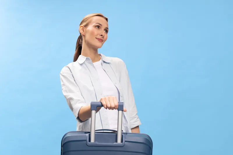 Young woman with suitcase stands against blue background looking away thoughtfully. Travel concept with casual outfit and luggage for vacation or business trip planning.