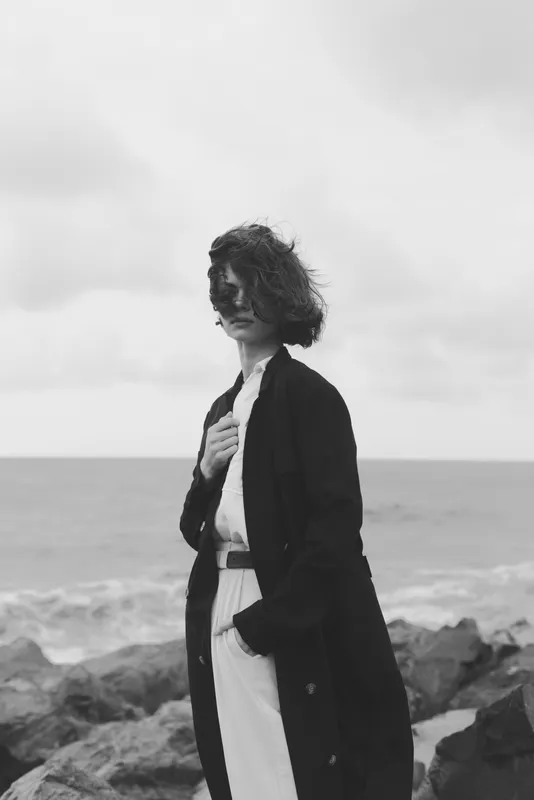 Woman in a long coat stands on a windy shore, monochrome scene by the ocean with rocks and waves, conveying timeless fashion and quiet strength