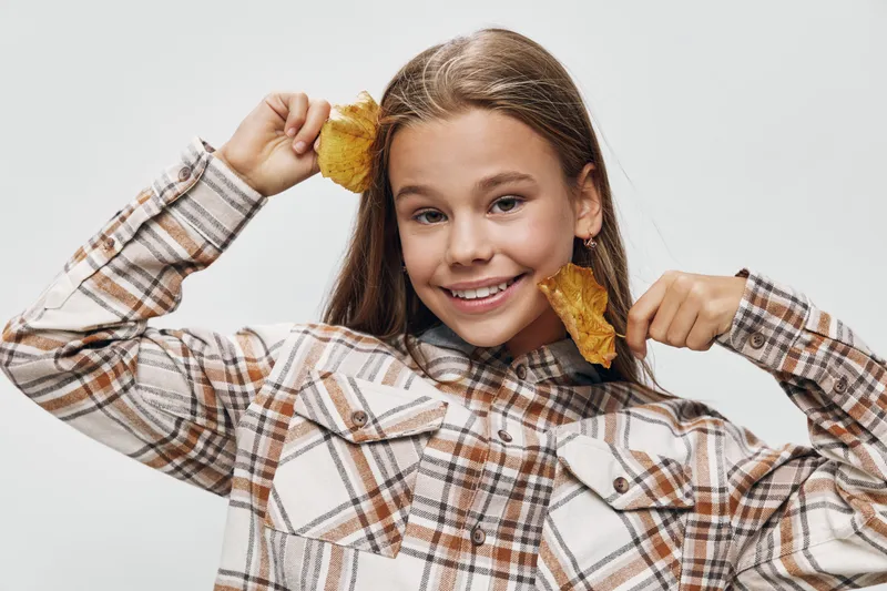 Girl holding yellow autumn leaves near face with playful smile wearing plaid shirt on light background. Child having fun with dry fall foliage for seasonal theme and childhood joy.