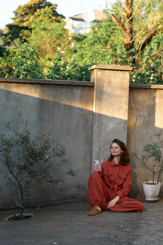 Young woman in a rust red outfit sits on the ground beside a concrete wall in a sunlit courtyard, relaxed and content, with soft plants and warm shadows creating a peaceful, candid outdoor moment