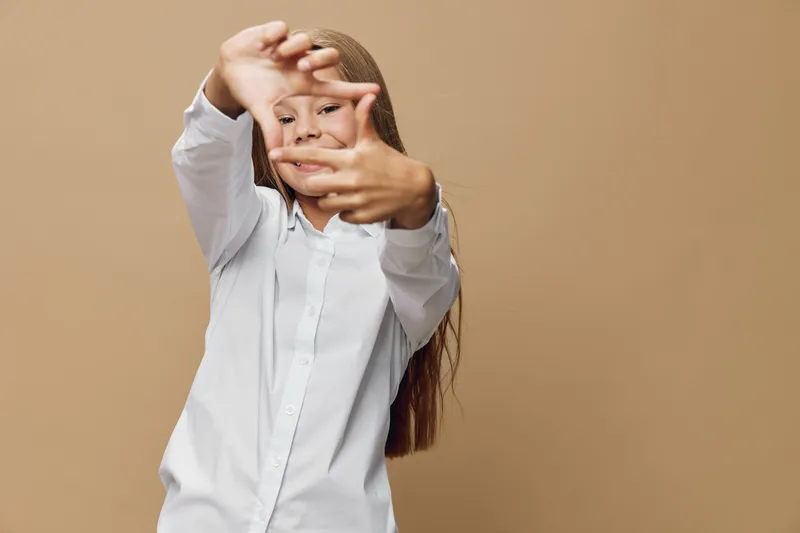 Girl in white shirt smiling and framing face with hands standing against beige background. Child expressing creativity and happiness with casual outfit and long hair.
