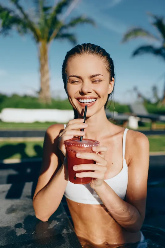 A smiling woman in a white bikini enjoys a refreshing smoothie by the pool on a sunny day, holding a straw. A lifestyle moment of leisure, wellness, and casual outdoor drinking.
