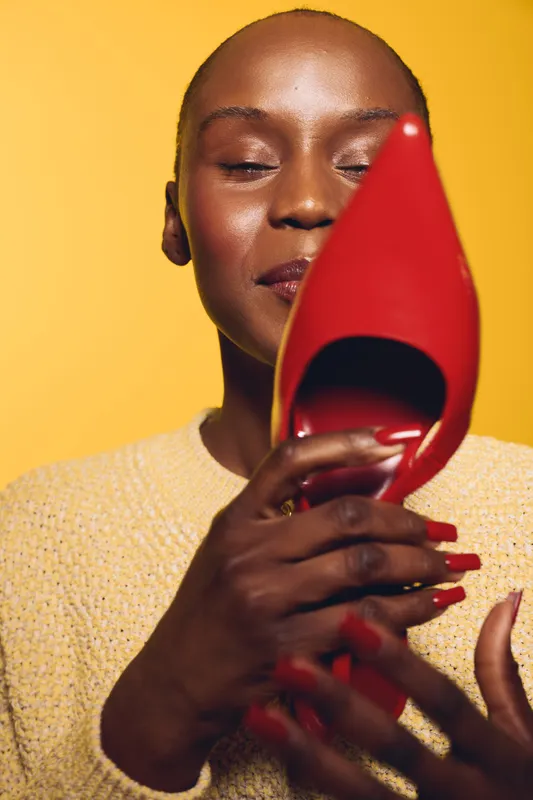 Close-up of a dark skinned African woman holding a red high heel shoe near her face with closed eyes. Concept of fashion, elegance, beauty, and style on yellow background.
