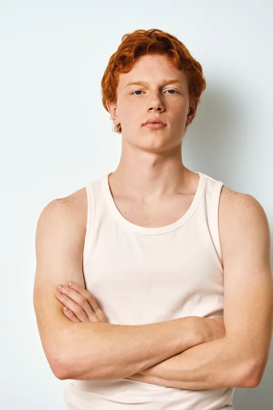 Young man with red hair and fair skin wearing white tank top standing with arms crossed looking confidently on white background studio. Casual style and natural expression portrait.
