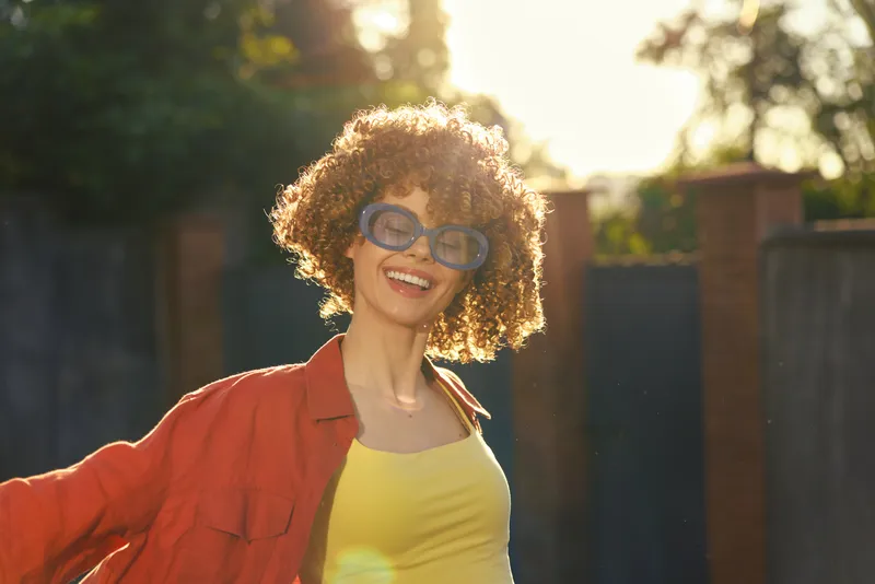 A smiling young woman with curly hair in a yellow top and red jacket enjoys an outdoor sunny day, exuding carefree energy and fashion-forward confidence