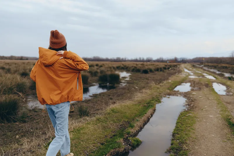 Woman with backpack, orange jacket, walking on a muddy road outdoors, seen from behind, near puddles on a field landscape, travel lifestyle concept, cloudy daylight