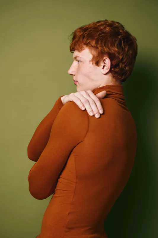 Young man with red hair in orange turtleneck sweater touching shoulder against green background, side profile portrait with calm expression and simple studio lighting.