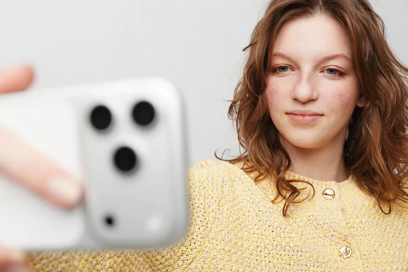 Young woman with wavy brown hair wearing yellow sweater takes selfie with smartphone on white background. Concept of social media, technology, communication, and casual lifestyle.