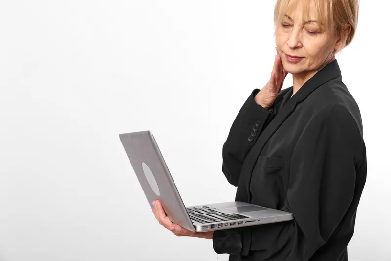elderly woman with laptop in office, hand on neck, showing pain, business health concept, studio white background, office worker worried expression, soft light