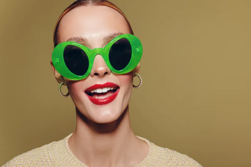 Smiling woman with bright red lipstick wearing large green sunglasses and hoop earrings on beige background. Concept of fashion, summer style, and confident beauty portrait.