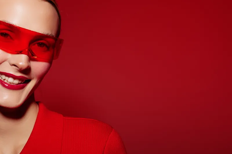 Smiling woman wearing red sunglasses and red sweater poses against red background. Close-up portrait with bright colors and confident expression. Concept of fashion and style.
