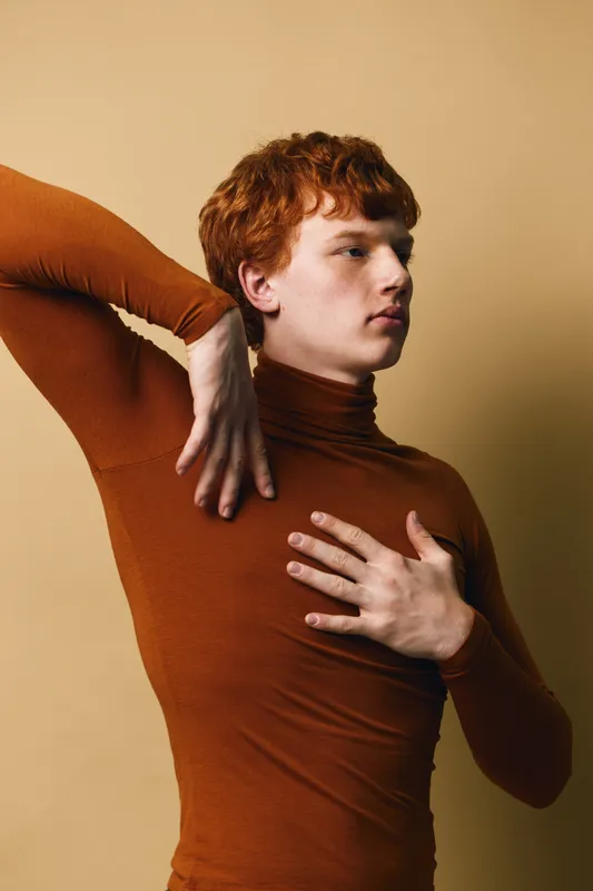 Young man with red hair in brown turtleneck poses with hand on chest and other bent behind neck against beige background. Studio portrait with thoughtful expression.