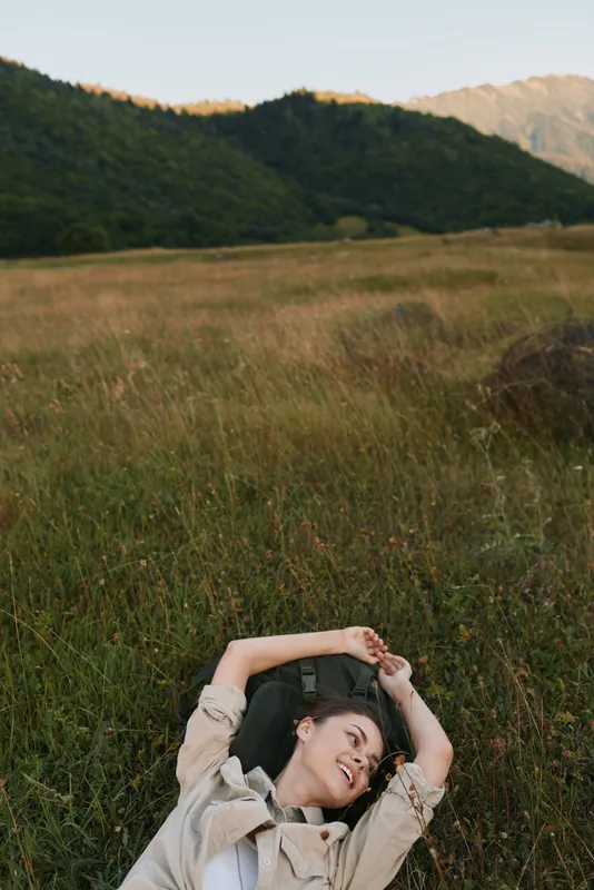 Young woman with short brown hair lies on tall grass in a natural field surrounded by hills, enjoying a peaceful moment with a relaxed and content expression.