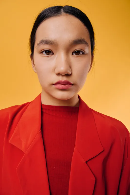 Portrait of young woman in red jacket and shirt with natural makeup against orange background. Serious expression, direct eye contact, minimalistic style for fashion or beauty concept.