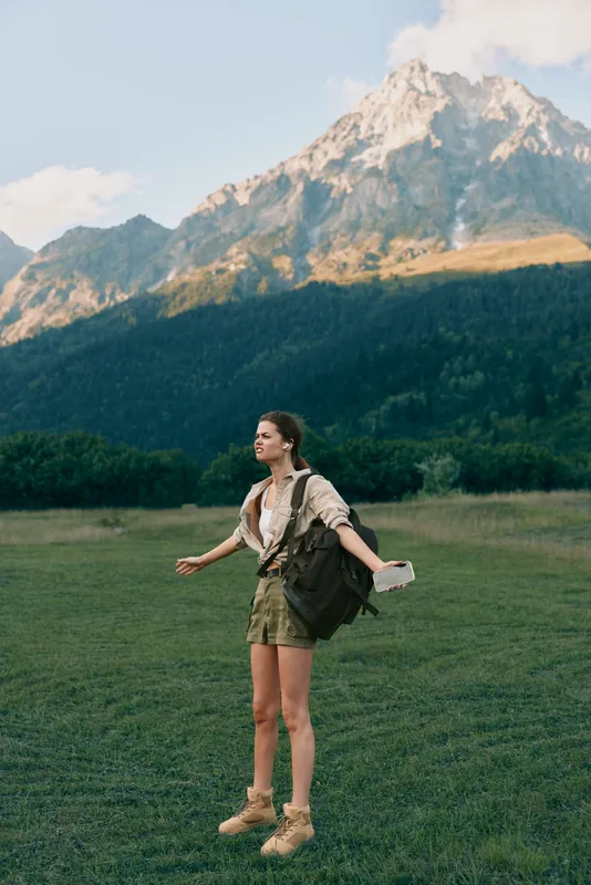 Cheerful woman outdoors, hiking through a grassy meadow with distant mountains, backpack strapped, sunlit scene, casual travel vibe, relaxed posture, natural colors, gentle breeze