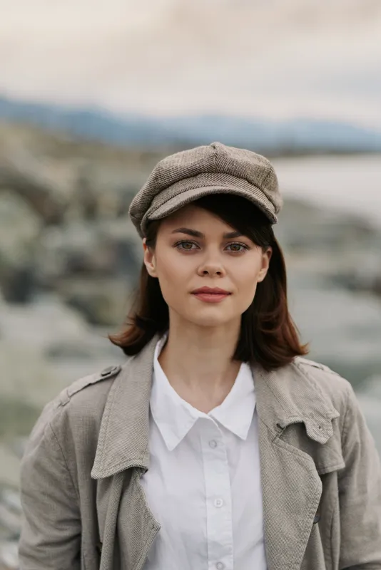 Young woman wearing a textured jacket and newsboy cap stands outdoors with blurred natural background. Portrait of casual style and autumn fashion concept.