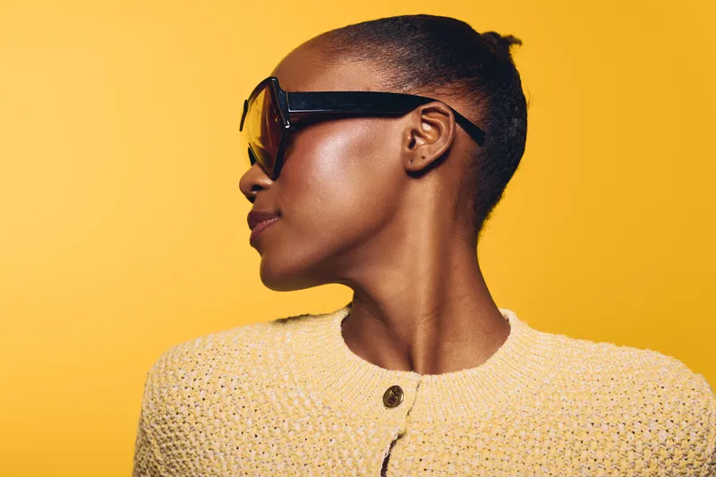Profile of a dark skinned African woman wearing large black sunglasses and a beige knitted sweater against yellow background. Concept for fashion, style, and summer accessories.