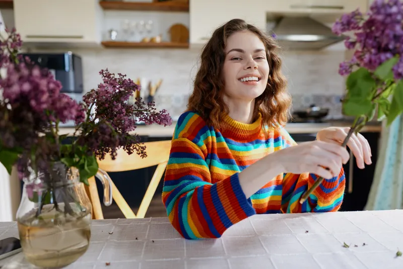 Cheerful woman indoors, colorful striped sweater, arranging flowers on a kitchen table, bright natural light, cozy home atmosphere, candid lifestyle moment, vibrant colors, calm smile