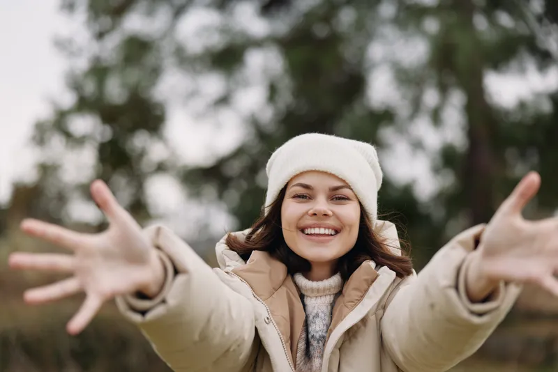 Smiling woman outdoors in a beige winter coat and white beanie with open arms, inviting and cheerful, posing in a park setting on a chilly day.