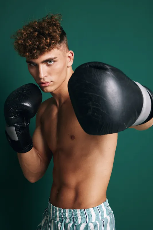 Young man with curly hair wearing boxing gloves and striped shorts training in fighting stance against green background for fitness and sport concept.