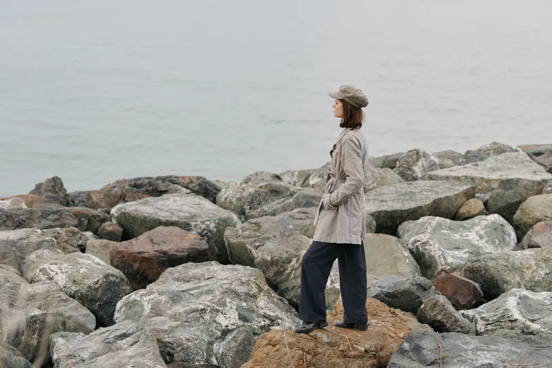 A woman in a trench coat and hat stands on rocky shore looking at the sea. Overcast sky and calm water create a peaceful, contemplative mood in nature setting.