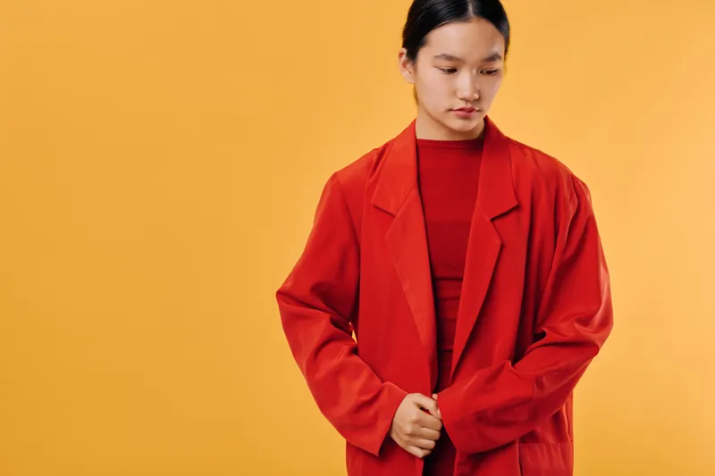 Young woman wearing oversized red jacket and matching top on orange background, looking down with thoughtful expression and hands clasped in front, studio portrait.
