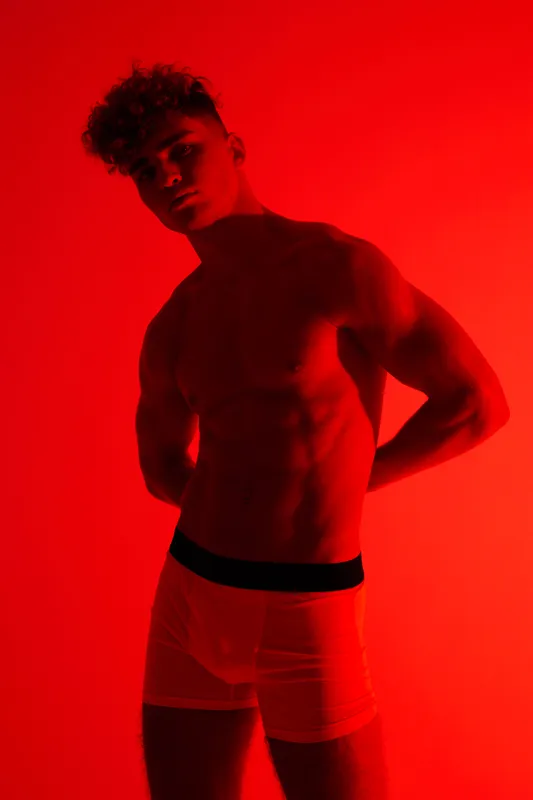 Young man with curly hair posing shirtless in tight boxer briefs, showing muscular body and shadows on bold red background. Fitness, confidence, and male silhouette highlight.