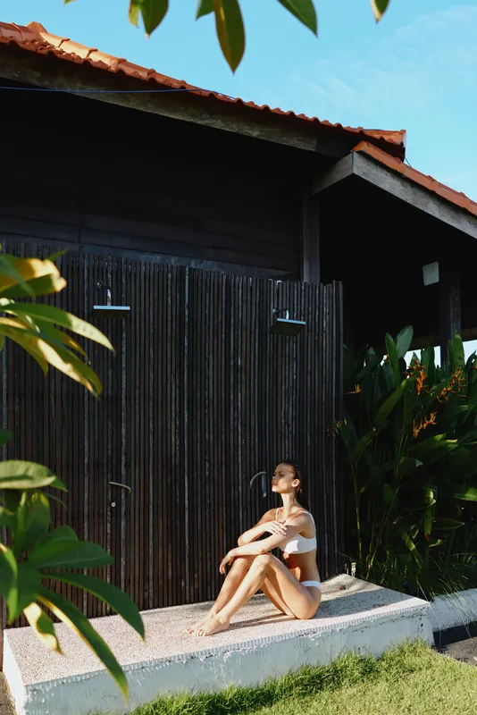 Young woman in white swimsuit sitting on the ground near outdoor wooden shower, relaxing in the sun, tropical plants around, concept of summer lifestyle and wellness.