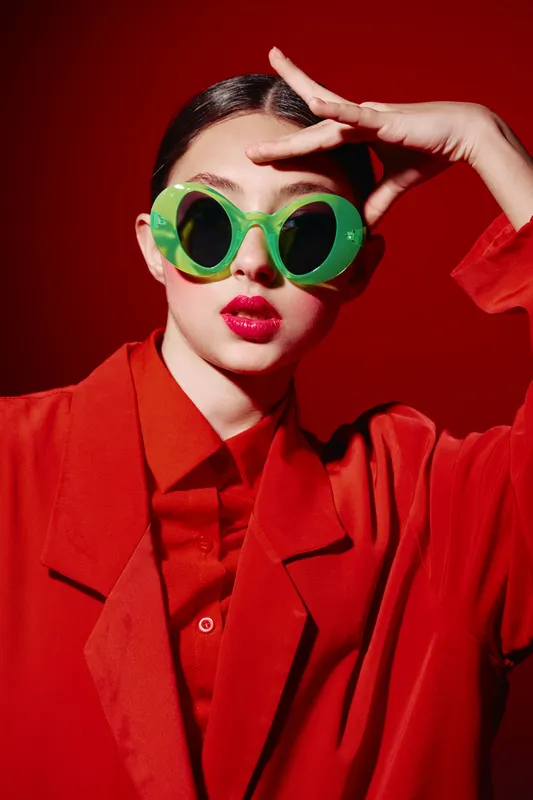 Stylish woman in a red outfit poses against a red background wearing large green sunglasses. Her bold makeup and confident gaze express contemporary fashion and beauty creativity.