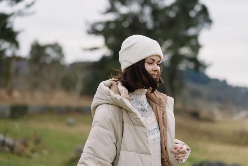 Woman in a beige puffer coat and white knit hat standing in a windy outdoor landscape, calm mood, soft natural light, seasonal style photography.