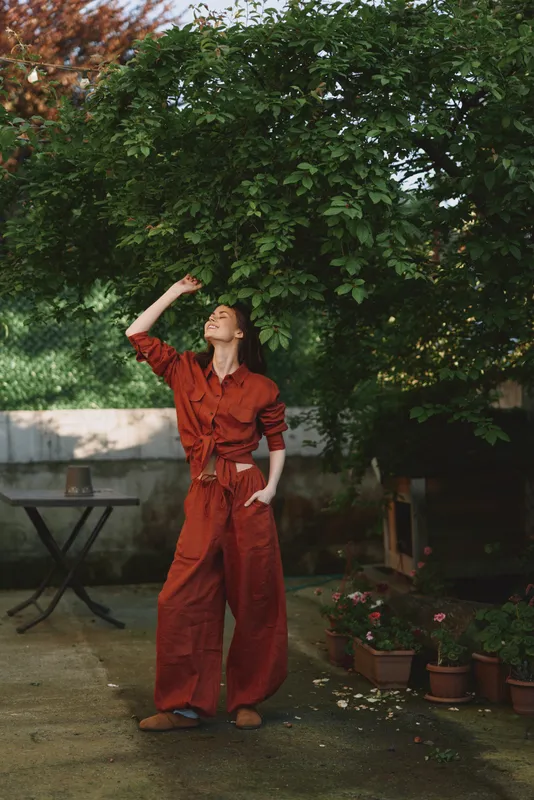 A woman in a red jumpsuit stands under a leafy tree on a patio, one hand on her hip and the other brushing hair, exuding relaxed confidence and playful style today