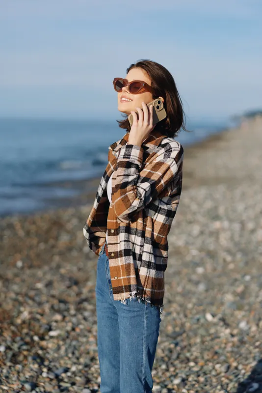Woman on a pebble beach in a plaid shirt and jeans, talking on a phone with sunglasses, enjoying a sunny seaside scene and a relaxed casual vibe by the shore.